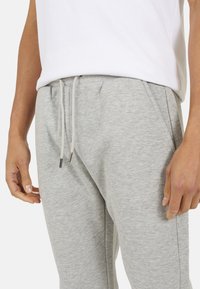 Persoon gekleed in lichtgrijze joggingbroek met touwtjes en een wit shirt, met ontspannen handen naast zich, tegen een effen achtergrond.