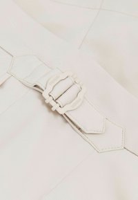 Nahaufnahme eines hellbeigen Stoffgürtels mit passender rechteckiger Schnalle, die mit „REISS“ graviert ist, an einem Kleidungsstück.