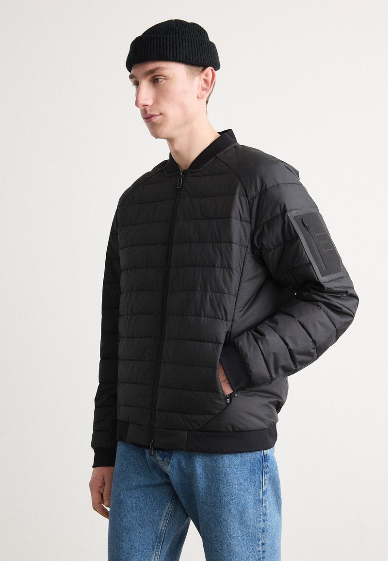 Veste bomber noire matelassée avec fermeture éclair sur le devant, poignets côtelés et un patch sur la partie supérieure de la manche. Associée à un jean bleu clair.