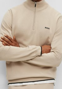 Beige halvmanschett sweatshirt med ribbad krage och manschetter. Har en liten svart logotyp och kontrasterande svarta detaljer på manschetterna och fållen.