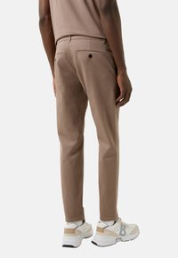 Bukser i lysebrunt stof, tætsiddende pasform, med to baglommer og en glat tekstur. Bruges med hvide og beige sneakers.