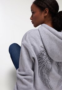 Felpa grigia con un grande design di ali nere sul retro. Presenta un cappuccio regolabile con lacci e un tessuto morbido e strutturato. Indossata con jeans blu.