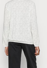 Eine Person trägt eine weiße Bluse mit langen Ärmeln, die kleine schwarze Punkte hat, und schwarze Hosen. Sie wird von hinten vor einem schlichten Hintergrund gezeigt.