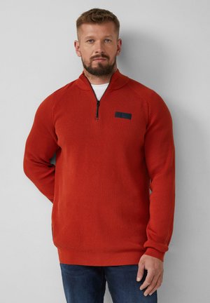 Roter Reißverschluss-Pullover mit strukturiertem Strickdesign, gerippten Bündchen und Saum sowie einem kleinen schwarzen Patch auf der linken Brust. Über einem weißen Shirt getragen.