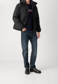 Musta toppatakki irrotettavalla hupulla, tikattu pinta, laivastonsininen neule vaakaraitoineen, siniset farkut ja mustat saappaat.