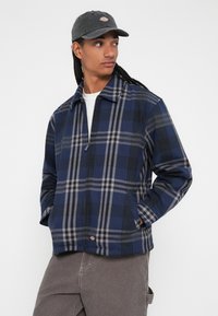 Casaco com fecho éclair em padrão xadrez nas cores azul marinho e cinza, feito de um tecido suave, com mangas longas, bolsos laterais e colarinho plano. Usado com um boné cinza.