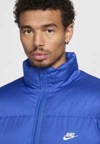 Mėlynas paminkštintas švarkas su aukšta kaklo linija, užtrauktuku ir Nike logotipu. Audinys turi glotnų paviršių ir blizgantį apdailą.