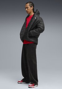 Sort pufjakke med quiltet design og hætte, rød skjorte under, løse mørke jeans og rød og sort sneakers.