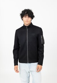 Zwarte zip-up jack met een staande kraag, rib gebreide manchetten en een ritssluiting op de linkermouw. Glad textuur met subtiele branding.