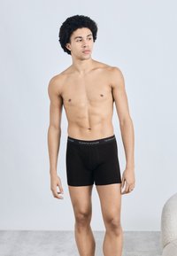 Sorte boksershorts med logo på elastiklinningen, lavet af blødt stof. Modellen står med armene afslappet og fremviser pasformen og designet.