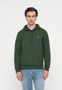 Hanorac verde dintr-un material texturat, având un model geometric subtil, cu glugă și șnur, și un mic logo brodat. Purtați cu blugi albaștri.