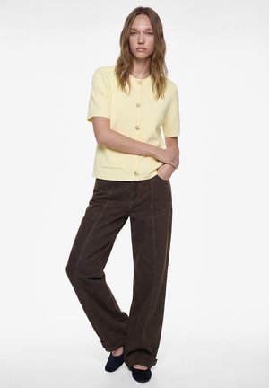 Vrouw draagt een lichtgeel kortärmelig shirt met knopen, donkerbruine wijde broekspijpen en zwarte schoenen, staand met één arm over de borst en één hand in de zak.