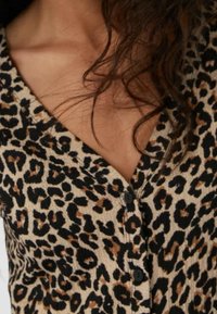 Mujer con una blusa beige de estampado de leopardo, con el cabello rizado oscuro y suelto parcialmente cubriendo la clavícula.