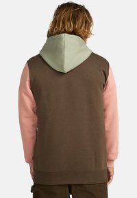 Sudadera con capucha que presenta un cuerpo marrón, mangas rosas y una capucha verde claro. Confeccionada en un material suave y texturizado con puños acanalados.