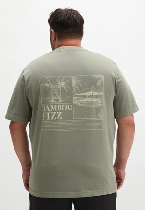 Mann trägt olivgrünes T-Shirt mit beiger Grafik eines tropischen Getränks, Palmen und dem Text "Bamboo Fizz" auf dem Rücken.
