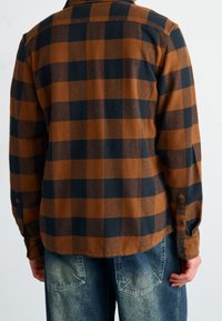 Camicia flanella a quadri marrone e nera, maniche lunghe, texture morbida, polsini con bottoni, orlo dritto, indossata con jeans in denim azzurro chiaro.