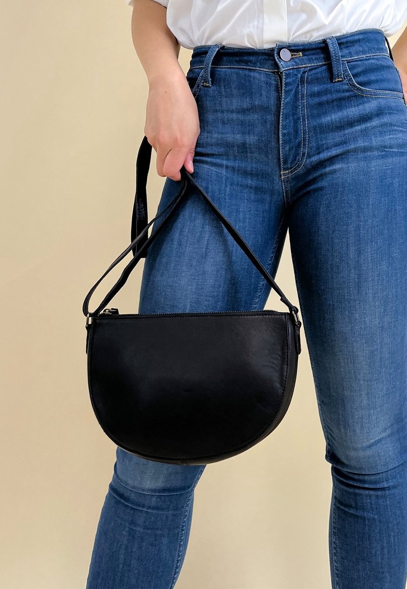 Borsa a tracolla in pelle nera con forma curva, chiusura a zip e tracolla regolabile, indossata da una persona in jeans blu.