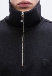 Sweat-shirt noir à demi-fermeture éclair avec un col haut, tissu texturé, fermeture éclair argentée et un patch logo carré noir sur la poitrine gauche.