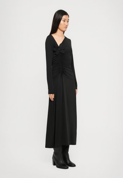 Hosszú ujjú fekete ruha, mellrészen ráncolással, virágos díszítéssel, áramló sziluettel és sima textúrával. Magassarkú csizmával viselve.