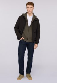 Svart jacka över en grön huvtröja, båda med dragkedjor. Vit skjorta med krage underneath. Mörkblå jeans och olivgröna sneakers fulländar outfiten.