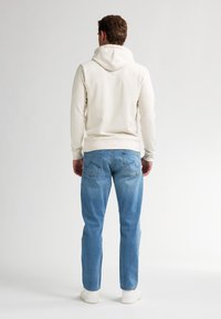 Cremefarbener Hoodie mit großer Kapuze auf der Rückseite, kombiniert mit hellblauen Jeans mit Gesäßtaschen und einem verwaschenen Finish. Slim-Fit-Design. Weiße Sneakers.