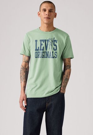 Homme avec des tatouages sur les deux bras portant un t-shirt Levi's Originals vert clair et un jean bleu foncé, debout devant un fond neutre.