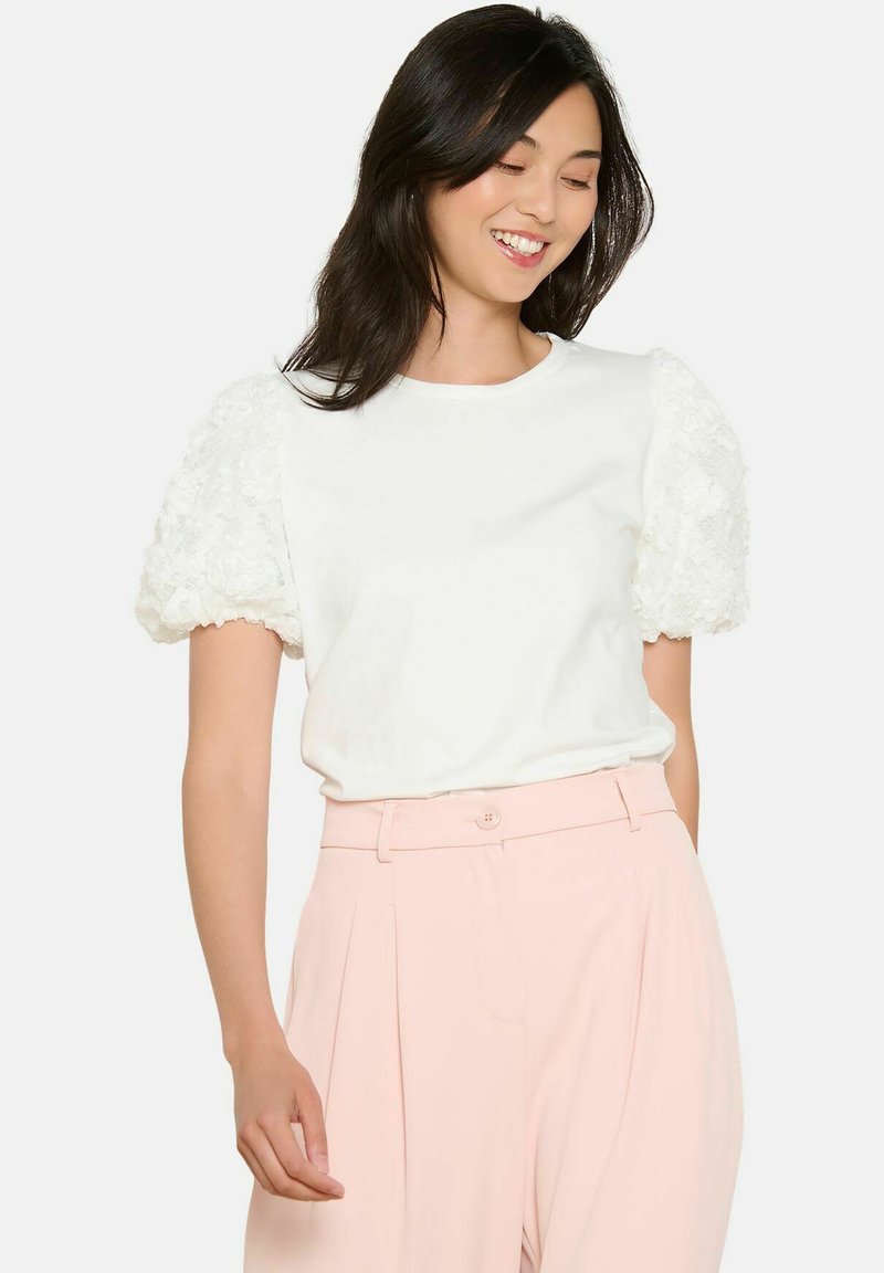 Balta īsām piedurknēm augšdaļa ar teksturētu mežģīņu pūkainām piedurknēm, apvienota ar gaiši rozā augsto jostu biksēm, kurām ir pogu aizdare.