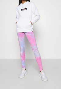 Žena v bílé mikině ellesse, růžovo-fialových legínách s tye-dye vzorem a bílých teniskách, stojí na světle neutrálním pozadí.