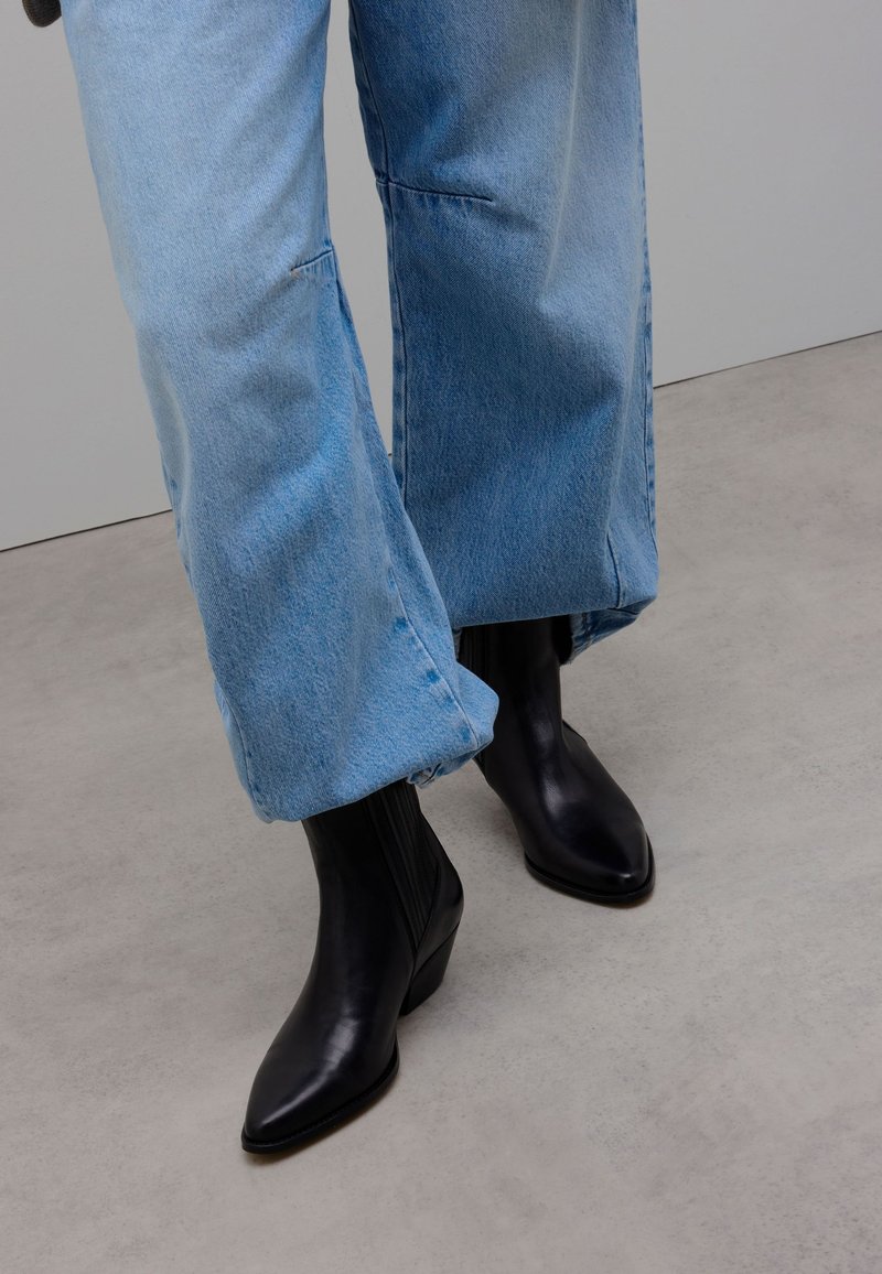 Personne portant un jean bleu clair à jambes larges et des bottines noires à bout pointu, debout sur un sol gris clair.