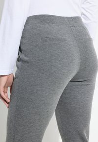 Primer plano de una persona que lleva pantalones gris ajustados con bolsillos traseros de ribete y una camisa blanca de manga larga.