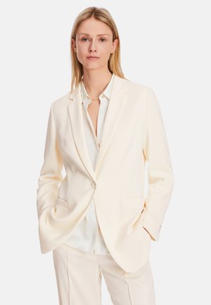 Cream-colored tailored blazer with a single button, notched lapels, and front pockets, worn over a light, collared shirt.