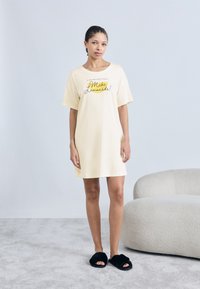 Φόρεμα σε ανοιχτό κίτρινο χρώμα τύπου t-shirt με άνετη εφαρμογή, διακοσμημένο με μαύρο κείμενο και γραφικά λεμονιών, συνδυασμένο με μαύρα αφράτα σανδάλια. Απαλή υφή υφάσματος.