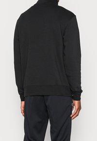 Man som bär en svart långärmad sweatshirt och svarta byxor, visad bakifrån mot en enkel ljus bakgrund.