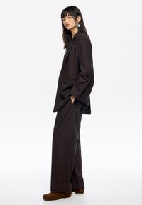 Abito a quadri marrone con giacca rilassata e pantaloni a gamba larga. Il materiale sembra texturizzato. Stivali alla caviglia marroni e accenti blu nei orecchini.