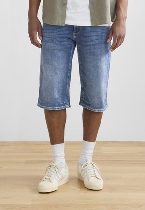 Man in blauwe denim knielange shorts, witte crew-sokken en witte sneakers staat op een houten vloer.