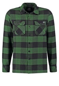 Chemise en flanelle à carreaux verts et noirs avec manches longues, boutons sur le devant, deux poches poitrine à rabats et un col, présentée sur fond uni.