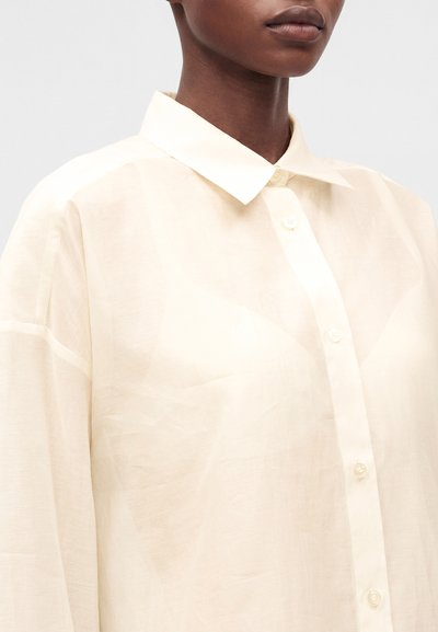 Personne portant une chemise ample beige clair à boutons avec un col pointu sur un fond uni, cadrée du nez vers le bas.