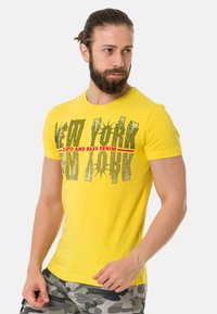 Gelbes Kurzarm-T-Shirt mit einem grafischen Aufdruck "NEW YORK" in Schwarz und Rot, aus Baumwollmischung gefertigt, getragen mit Camouflage-Shorts.