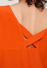 Orange top med V-rygdesign, der har krydsende stropper, glat tekstur og en enkel, udekoreret stil.