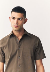 Braunes kurzärmeliges Hemd mit strukturiertem Muster, Brusttasche und Knopfleiste. Verfügt über einen klassischen Kragen und blaue Knöpfe.