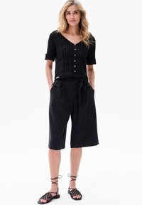 Zwarte gebreide cropped top met korte mouwen en knopen aan de voorkant, gecombineerd met hoge zwarte wijde shorts met een strikceintuur. Zwarte verstelbare sandalen.