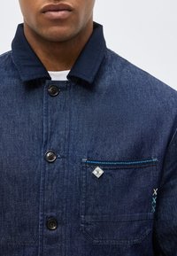 Άνδρας που φοράει σκούρο μπλε denim μπουφάν με μαύρα κουμπιά, τσέπη στο στήθος με μπλε ραφή και ένα μικρό λευκό λογότυπο.