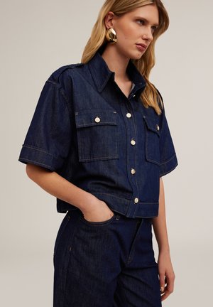 Dark blue denim short-sleeve shirt with two chest pockets and silver buttons. Features a collar and slightly cropped design.