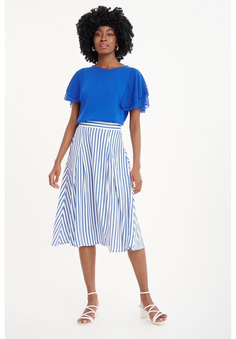 Top azul texturizado con mangas transparentes con volantes, combinado con una falda blanca con rayas verticales azules y una cinturilla ajustada. Zapatos blancos de tiras.