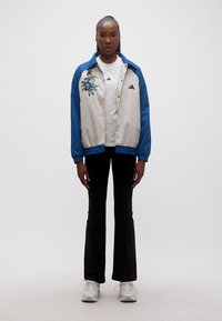 Γυναίκα που στέκεται φορώντας μπλε και μπεζ μπουφάν με σχέδιο πουλιού, λευκό μπλουζάκι, μαύρο παντελόνι και λευκά αθλητικά παπούτσια σε λευκό φόντο.