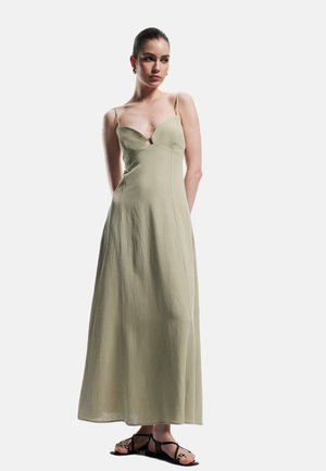 Vestido maxi sin mangas, de color verde claro, con escote en V y un ligero descenso en el escote. Confeccionado en una tela suave con un diseño fluido. Sandalias simples complementan el look.