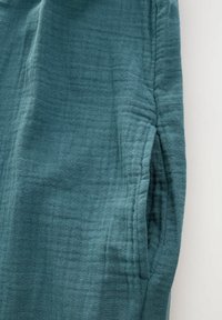 Tejido en color verde azulado con una textura de tejido relajado. Presenta un detalle de bolsillo oculto y un suave drapeado, que resalta un diseño desenfadado.