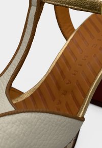 Κρεμ δερμάτινο σανδάλι με χρυσή λωρίδα διακόσμησης, που διαθέτει καφέ λαστιχένια σόλα με υφή και λεπτομερείς ραφές κατά μήκος των ακρών.