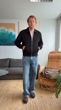 Giacca nera con zip e due tasche frontali sopra una camicia chiara, abbinata a jeans blu e scarpe scure. In piedi in una stanza con piante.