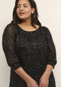 Haut noir à paillettes avec une texture lisse, un col rond et des manches bouffantes trois-quarts. Le tissu capte la lumière, créant un intérêt visuel.
